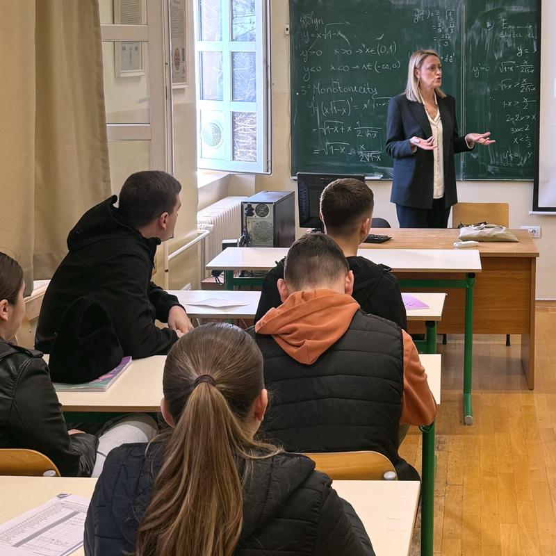 Facing the front of a classroom, four students are engaged listening to their teacher who is stood at the front