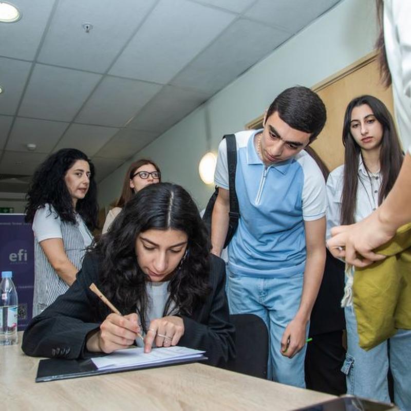 Students looking intently at a female who is writing on a piece of paper at a desk