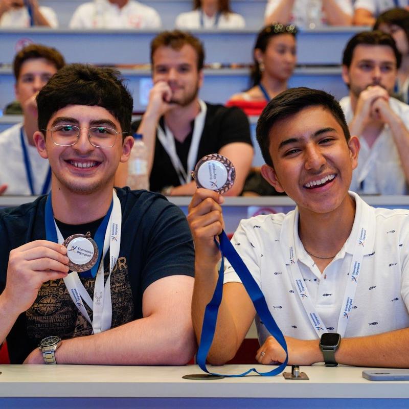 Two students smiling and happy, holding up their medals to the camera at the Economics Olympiad