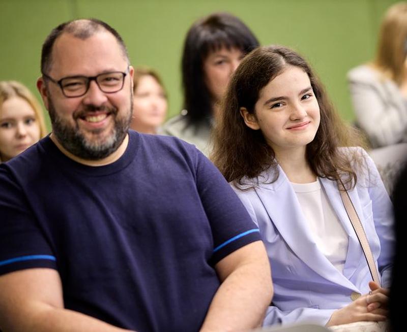 A male wearing glasses and a female next to him, sat down, smiling and looking directly at the camera