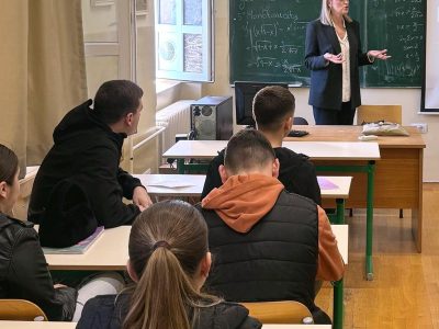 Facing the front of a classroom, four students are engaged listening to their teacher who is stood at the front