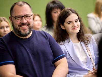 A male wearing glasses and a female next to him, sat down, smiling and looking directly at the camera